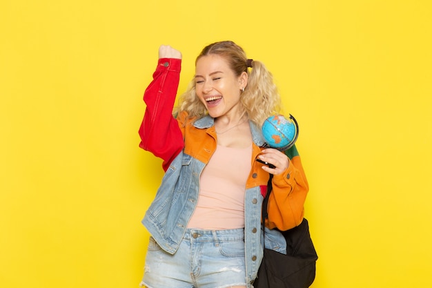 Studentin jung in der modernen Kleidung, die kleinen Globus mit glücklichem Gesicht auf Gelb hält