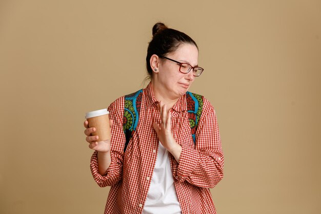 Studentin junge Frau in legerer Kleidung mit Brille und Rucksack mit Kaffeetasse, die mit angewidertem Gesichtsausdruck schaut und Stoppschild mit offener Hand über braunem Hintergrund macht