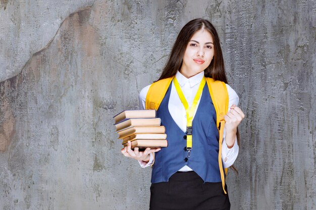 Studentin mit gelbem Rucksack und Büchern, die über Grau stehen. Foto in hoher Qualität