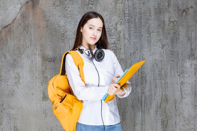 Studentin mit gelbem Rucksack und Kopfhörern, die mit Buch stehen. Foto in hoher Qualität
