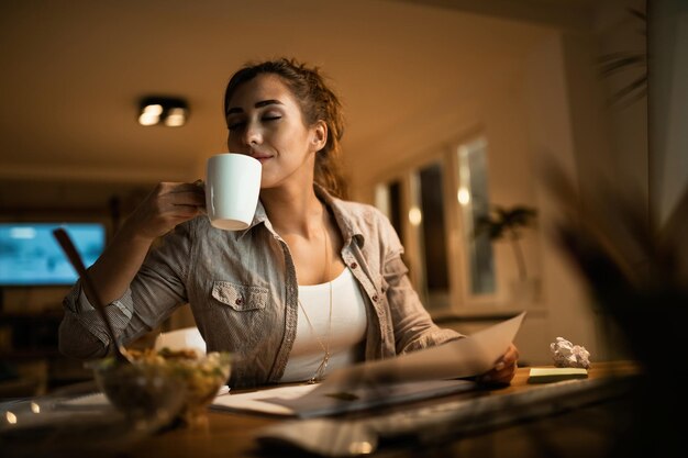 Studentin mit geschlossenen Augen, die den Geruch von frischem Kaffee genießt, während sie abends zu Hause lernt