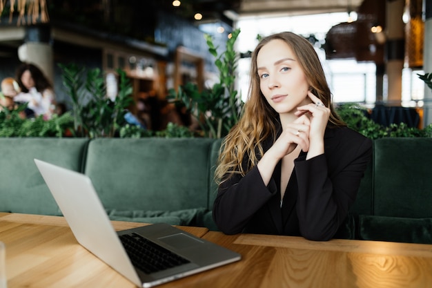 Studentin mit niedlichem Lächeln, das etwas auf netbook tastet, während sie sich nach Vorlesungen in der Universität entspannt, schöne glückliche Frau, die während der Kaffeepause in der Café-Bar am Laptop arbeitet