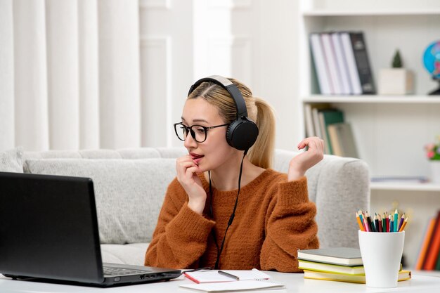 Studentin online, junges süßes Mädchen mit Brille und orangefarbenem Pullover, das am Computer beißendem Finger studiert