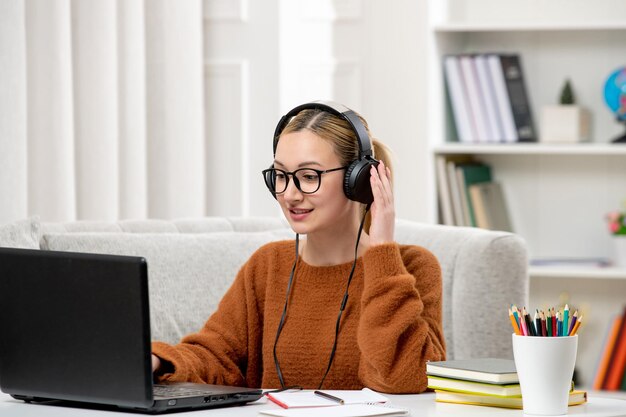 Studentin online, junges süßes Mädchen mit Brille und orangefarbenem Pullover, das am Computer mit Kopfhörern studiert