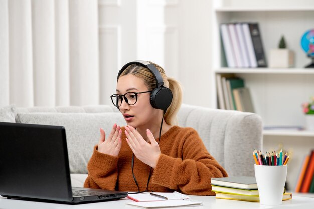 Studentin online, junges süßes Mädchen mit Brille und orangefarbenem Pullover, das am Computer studiert, schockiert