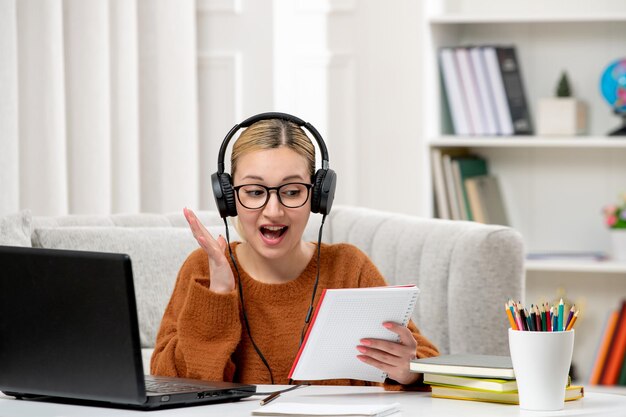 Studentin online süßes Mädchen in Brille und Pullover, das am Computer studiert, aufgeregt über ihre Notizen