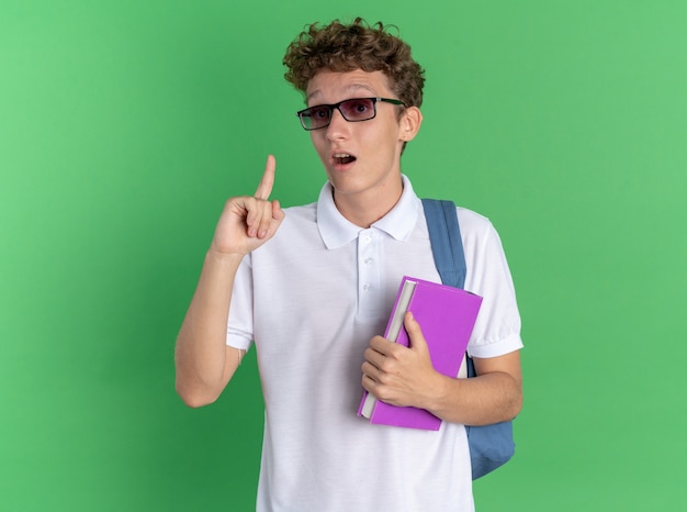 Studentischer Kerl in Freizeitkleidung mit Brille mit Rucksack, der ein Notizbuch hält, das den Zeigefinger zeigt, der eine neue Idee hat, die glücklich und aufgeregt auf grünem Hintergrund steht