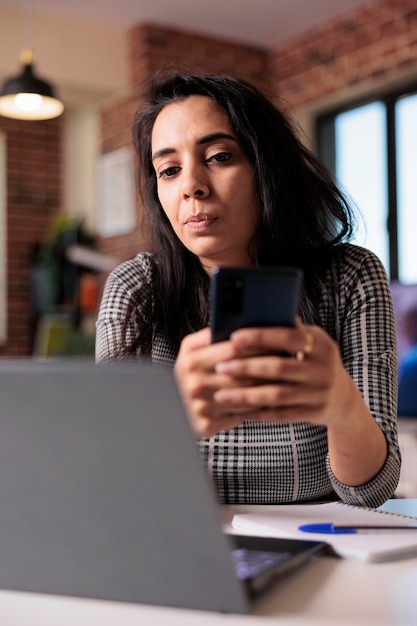 Studentischer mitarbeiter, der auf dem smartphone im internet surft und die social-media-app verwendet, um sms-nachrichten in der arbeitspause zu hause zu senden. arbeiten an remote-jobs mit laptop und mobiltelefon, suche nach online-informationen.