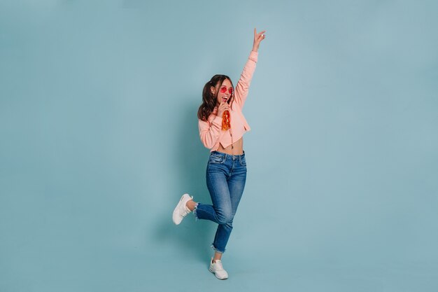 Studio-Foto der vollen Länge des jungen attraktiven Mädchens, das rosa Pullover und rosa runde Gläser trägt, die tanzen und Saft über isolierter Wand trinken