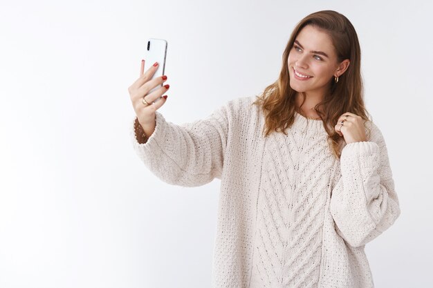 Studio Shot Glamour moderne attraktive Frau trägt einen stylischen, lockeren, gemütlichen Pullover, streckt den Arm aus und hält den Smartphone-Kippkopf posiert lächelnd auf dem Display und nimmt süßen Selfie-Post online, weißer Hintergrund