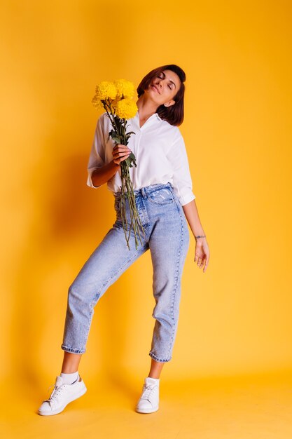 Studioaufnahme auf gelbem Hintergrund Glückliches kurzes Haar der kaukasischen Frau, das weißes Hemd der Freizeitkleidung und Jeanshosen hält, die Strauß der gelben Astern halten
