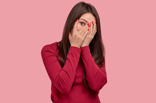 Studioaufnahme der attraktiven Frau schaut durch Finger, bedeckt Gesicht mit Händen, hat rote Maniküre, trägt Freizeitkleidung, posiert vor rosa Hintergrund