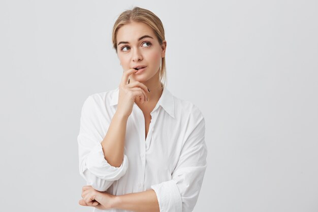 Studioaufnahme der gerissenen schönen Frau mit dem geraden blonden Haar, das beiseite hält Hand unter Kinn hält, um kniffligen Plan zu verwirklichen.