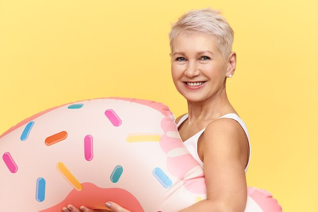 Studioaufnahme der glücklichen positiven reifen kaukasischen Frau mit Pixie-Frisur, die aufblasbaren Kreis in Form des rosa Donuts trägt, während der Sommerferien im See oder im Meer schwimmt und freudig lächelt