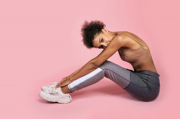 Studioaufnahme der hübschen schwarzen Frau mit afrikanischer Frisur in der stilvollen Sportbekleidung, die auf rosa Hintergrund sitzt.