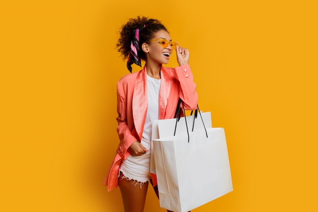 Studioaufnahme der hübschen schwarzen Frau mit der weißen Einkaufstasche, die über gelbem Hintergrund steht. Modischer Look im trendigen Frühling.