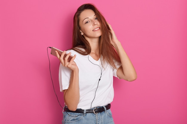 Studioaufnahme der jungen Frau, die Musik mit Kopfhörern hört, Smartphone in Händen hält, Kamera betrachtet, zu Hause entspannt