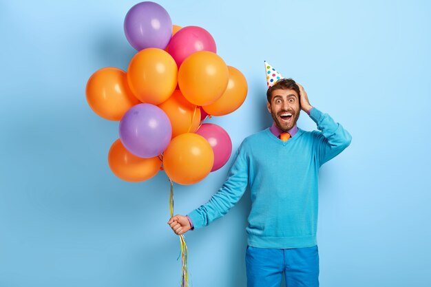 Studioaufnahme des aufgeregten Kerls mit Geburtstagshut und Luftballons, die im blauen Pullover aufwerfen