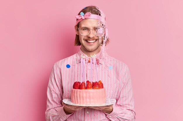 Studioaufnahme des glücklichen Mannes feiert Geburtstag hält leckeren Erdbeerkuchen trifft Gäste, die in festlichen Kleidern über rosa Hintergrund isoliert sind