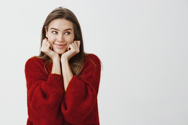 Studioaufnahme des glücklichen niedlichen kaukasischen weiblichen Mitarbeiters im stilvollen losen Pullover, der sich auf Hände stützt und mit neugierigem fröhlichem Lächeln beiseite schaut, in guter Stimmung ist und auf ihre Bestellung im Restaurant wartet