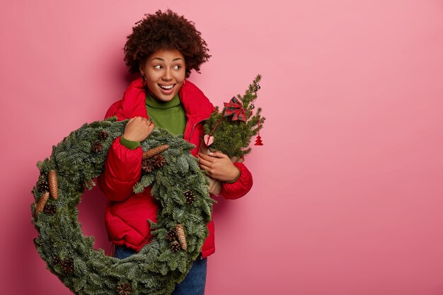 Studioaufnahme des schönen dunkelhäutigen Modells trägt Weihnachtskranz und geschmückten Tannenbaum, hat festliche Stimmung, trägt rote Oberbekleidung, isoliert auf rosa Hintergrund
