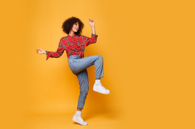 Studioaufnahme des schwarzen Mädchens, das mit glücklichem Gesichtsausdruck auf hellem orange Hintergrund springt. Trägt Jeans, weiße Turnschuhe und ein rotes Hemd.