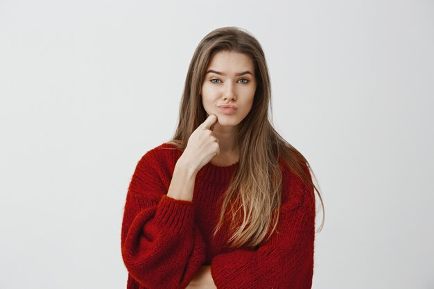 Studioaufnahme einer enttäuschten kaukasischen Frau in einem roten, losen Pullover, der einen Finger am Kinn hält und schmollt, unzufrieden und zweifelhaft aussieht und etwas Uninteressantes sieht