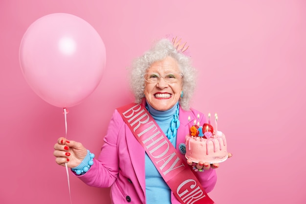 Studioaufnahme einer glücklichen, faltigen Rentnerin mit hellem Make-up, die zahnig lächelt, hält festlichen Kuchen mit brennenden Kerzen hat festliche Stimmung trägt aufgeblasenen Ballon