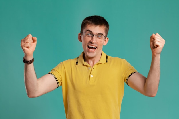 Studioaufnahme einer interessanten jungen Person in gelbem Freizeit-T-Shirt, Brille und schwarzen Uhren, die glücklich aussieht, während sie vor blauem Hintergrund posiert. Stylischer Haarschnitt. Konzept der aufrichtigen Emotionen. Leerzeichen kopieren
