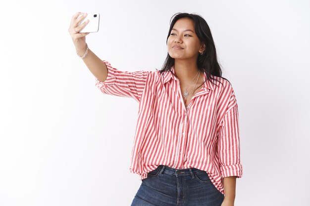 Studioaufnahme einer stylischen jungen, süßen asiatischen Frau, die Selfie macht, um Follower im Internet mit neuer trendiger Bluse zu beeindrucken, die die Hand mit dem Smartphone ausstreckt, das auf den Handy-Bildschirm schaut und fotografiert