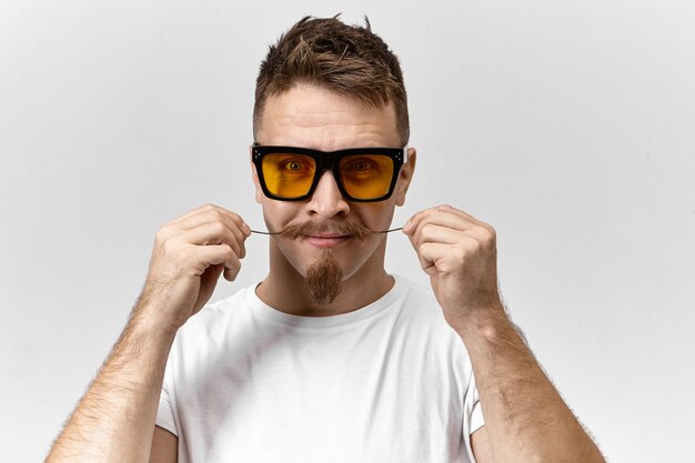 Studioaufnahme eines attraktiven jungen brünetten Mannes, der eine stilvolle gelb getönte Sonnenbrille und ein lässiges T-Shirt trägt, das sich um seinen Lenkerschnurrbart kümmert, sich mit Wachs kräuselt und sich auf ein Date vorbereitet