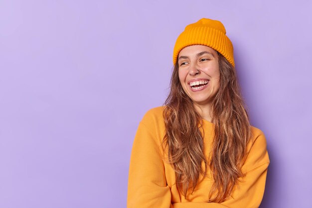 Studioaufnahme eines glücklichen Teenager-Mädchens mit einem zahnigen Lächeln, das freudig lacht, hat ein Piercing in der Nase, trägt einen orangefarbenen Hut und ein Sweatshirt, isoliert auf violettem Leerraum für Ihre Werbeinhalte