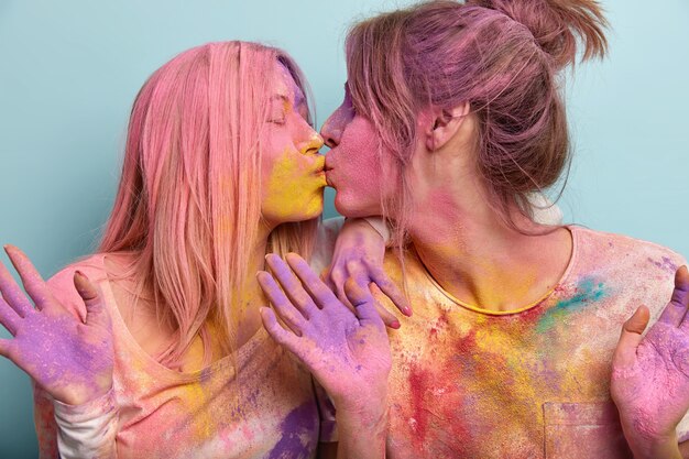 Studioaufnahme von erfreuten zwei europäischen jungen Frauen, die sich küssen, verschmiert mit buntem Holi-Pulver