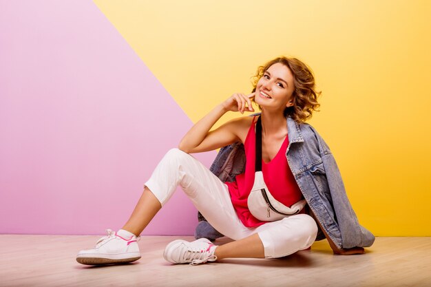 Studiobild der lächelnden brünetten reizenden Frau, die stilvolles sportliches Outfit und Jeansjacke sitzt, die auf dem Boden sitzen.