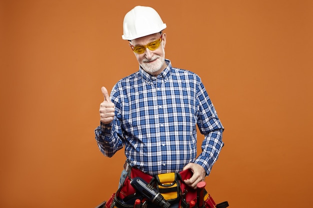 Studiobild des fröhlichen glücklichen älteren älteren bärtigen Reparaturmanns, der Helm trägt