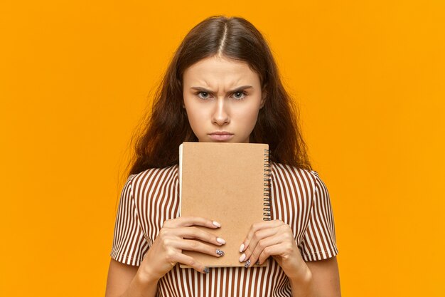 Studiobild des mürrischen weiblichen Teenagers mit dem langen losen Haar, das Tagebuch an ihrem Gesicht hält