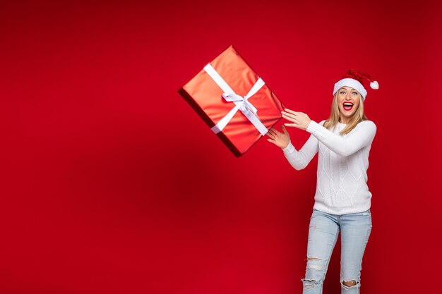 Studiofoto einer aufgeregten blonden Dame in Weihnachtsmütze, die mit weit geöffnetem Mund eine Geschenkbox in die Luft wirft. Platz kopieren