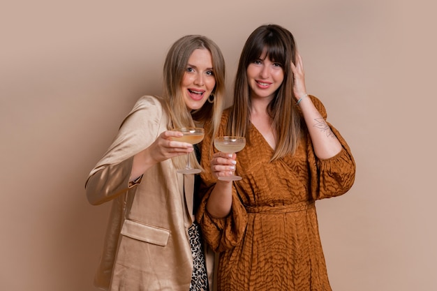 Studiofoto von zwei eleganten Frauen im stilvollen Herbst autfit, die mit einem Glas Champagner über beige Wand posieren