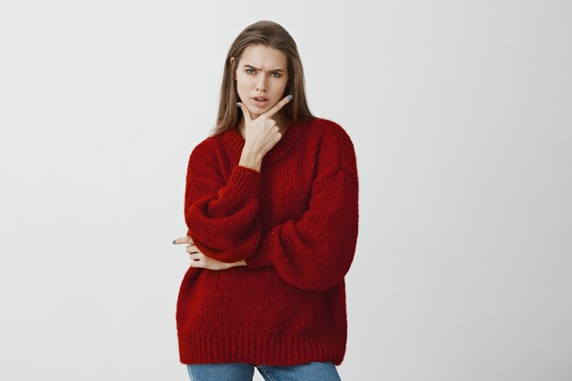 Studioporträt der unruhigen zweifelhaften attraktiven Frau im stilvollen roten losen Pullover, die Waffengeste auf Kinn hält und die Stirn runzelt, sich misstrauisch und frustriert fühlt und steht