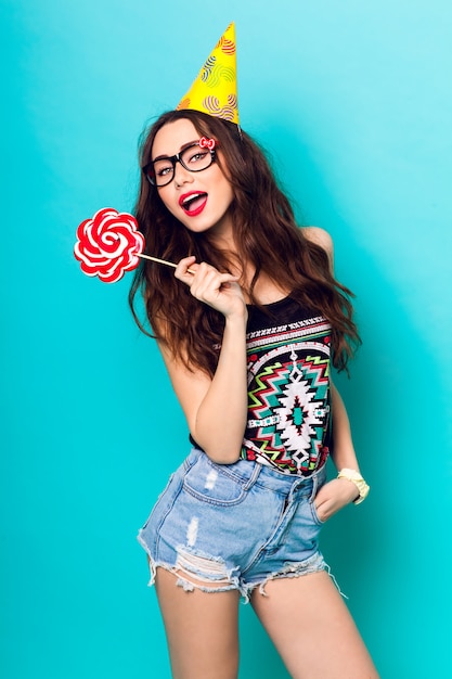 Studioporträt des jungen lustigen Mode-Mädchens, das auf blauem Wandhintergrund im Sommerart-Outfit mit rosa Lutscher aufwirft, der einen Papierhut und niedliche Brille trägt.