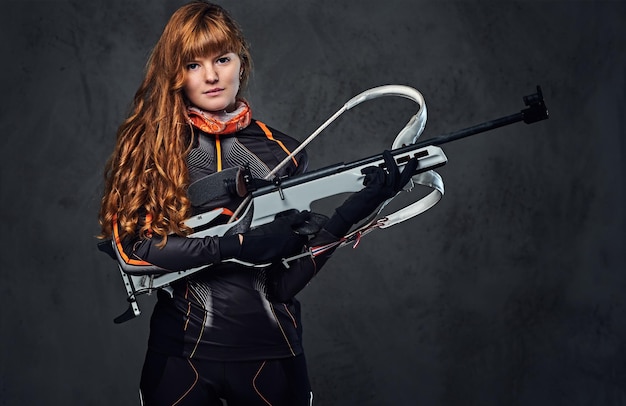 Studioporträt einer rothaarigen Biathlon-Meisterin hält eine Waffe auf grauem Hintergrund.