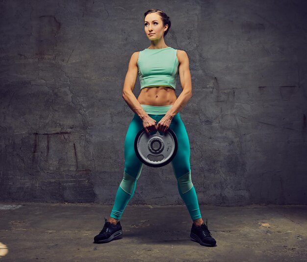 Studioporträt einer sportlichen Frau in azurblauer Sportbekleidung, die mit Langhantelgewicht auf grauem Hintergrund trainiert.