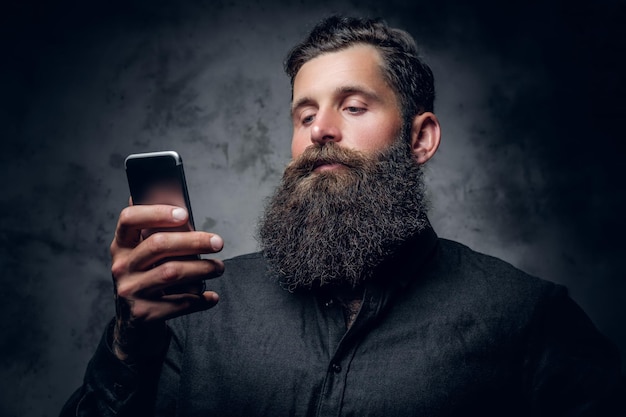 Studioporträt eines bärtigen Mannes hält ein Smartphone auf grauem Hintergrund.