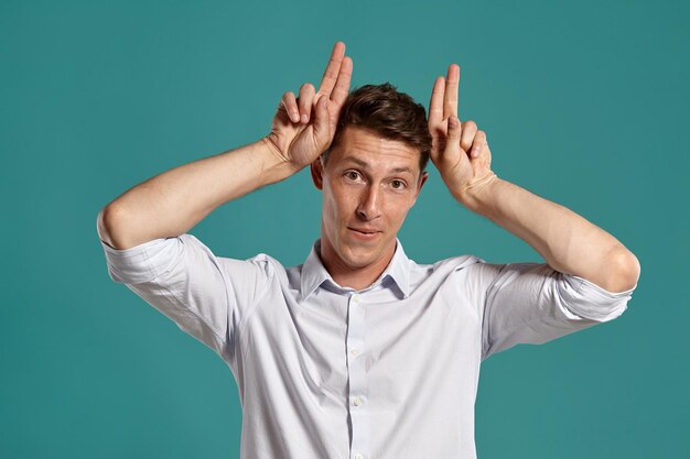 Studioporträt eines brünetten jungen Kerls in einem klassischen weißen Hemd, der vor blauem Hintergrund posiert, als wäre er eine Kuh. Stylischer Haarschnitt. Aufrichtiges Gefühlskonzept. Platz kopieren.