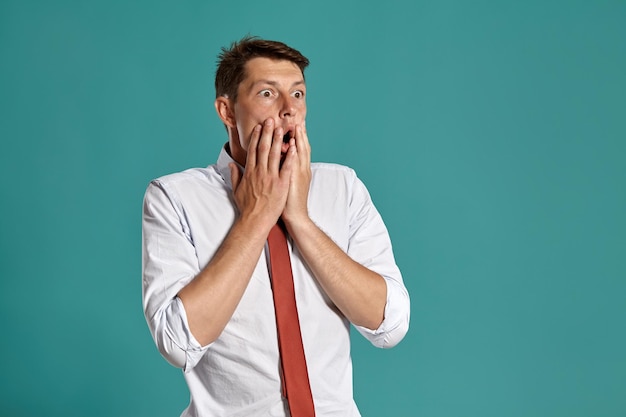 Studioporträt eines eleganten jungen Mannes in klassischem weißem Hemd und roter Krawatte, der schockiert aussieht, während er vor blauem Hintergrund posiert. Stylischer Haarschnitt. Aufrichtiges Gefühlskonzept. Platz kopieren.