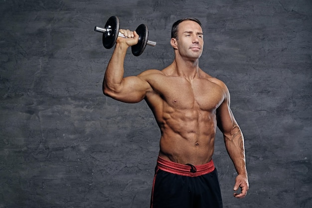 Studioporträt eines hemdlosen muskulösen Mannes mittleren Alters hält eine Hantel isoliert auf grauem Hintergrund.