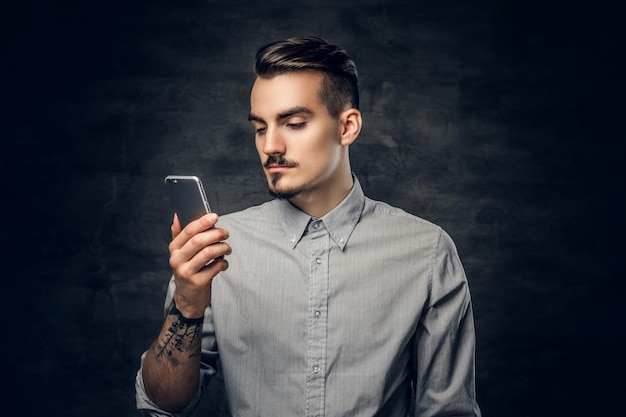 Studioporträt eines hübschen bärtigen Hipster-Männchens mit einer Tätowierung auf seinem Arm mit einem Smartphone.