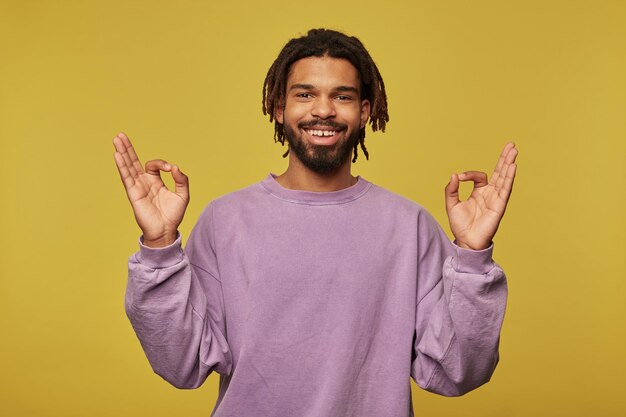 Kostenloses Foto studioporträt eines jungen afroamerikanischen mannes, der vor studiohintergrund posiert und mudra-zeichen zeigt, während er meditiert, lächelt breit und fühlt sich glücklich