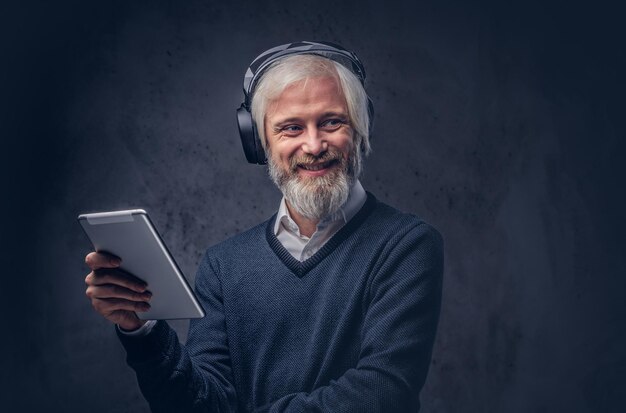 Studioportrait eines hübschen älteren Mannes, der ein Tablet mit Kopfhörern über einem dunklen Hintergrund verwendet.