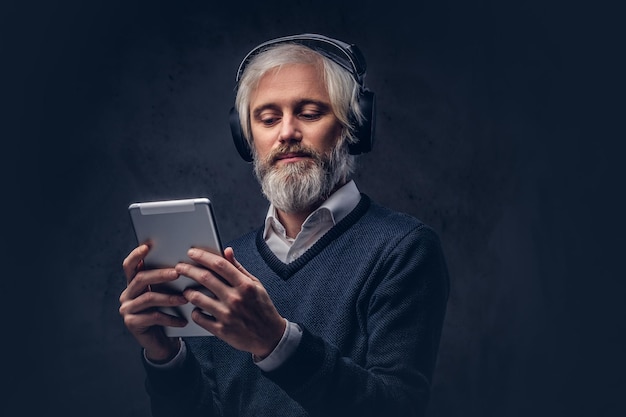 Studioportrait eines hübschen älteren Mannes, der ein Tablet mit Kopfhörern über einem dunklen Hintergrund verwendet.